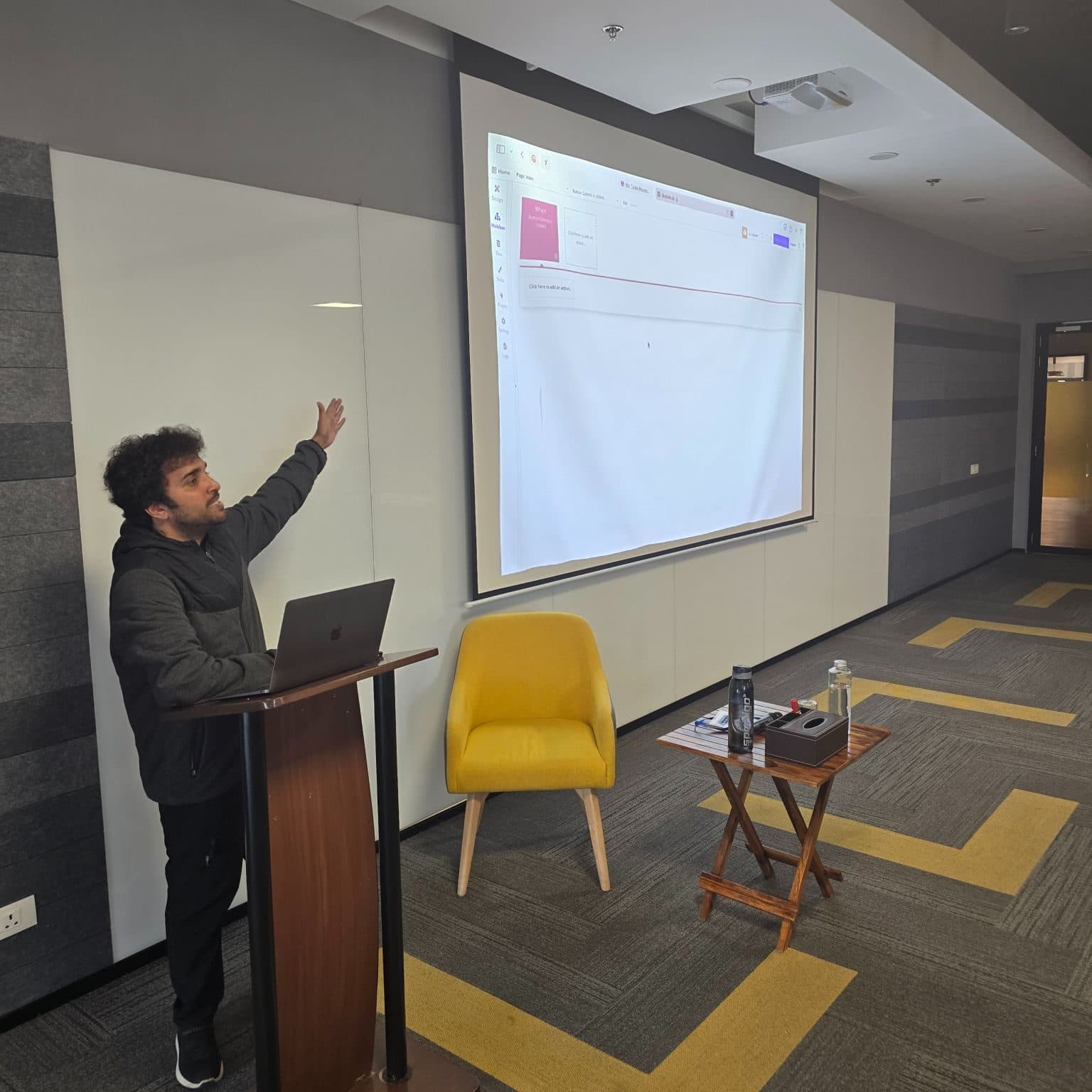 Adi teaching an AI workshop from a podium, pointing at a projected no-code editor.